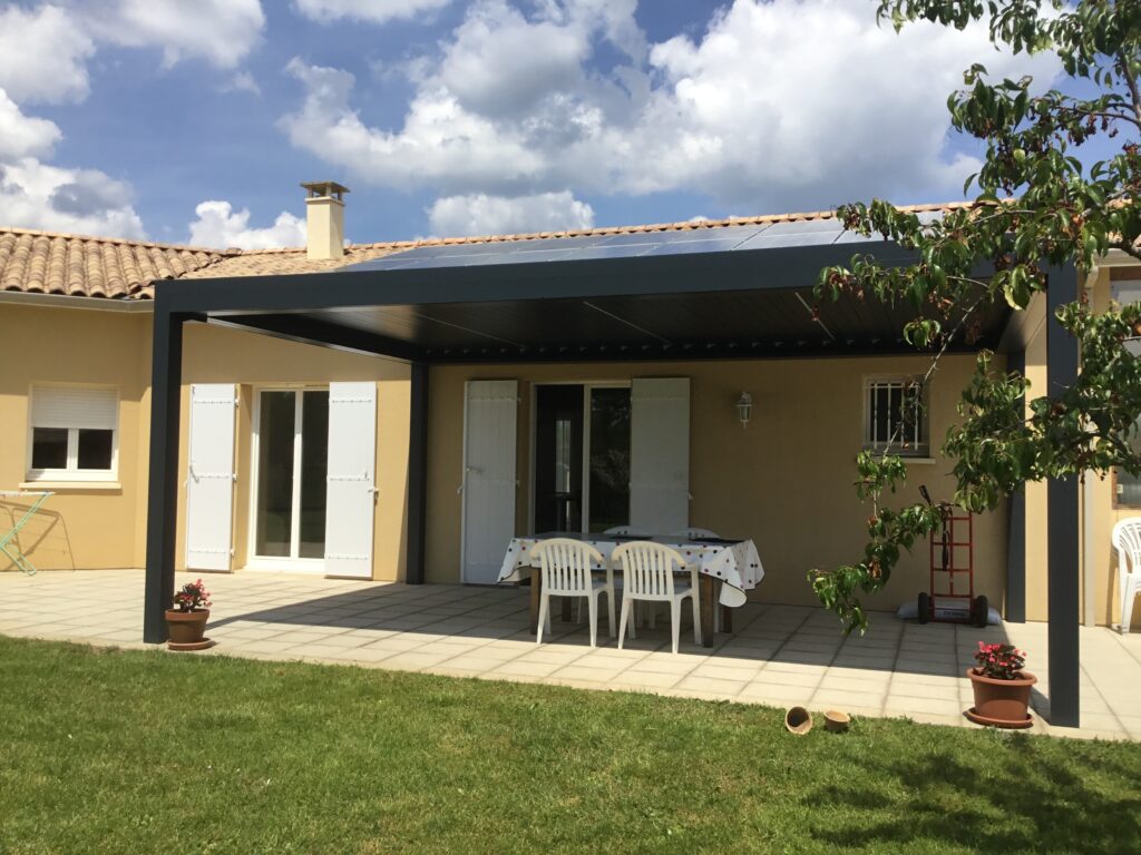 Pergola bioclimatique noire avec panneaux solaires sur une terrasse carrelée devant une maison avec jardin.