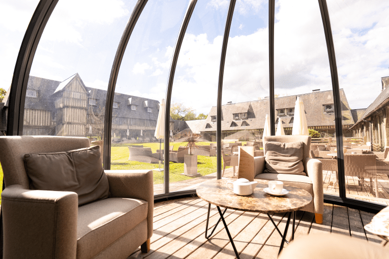 Intérieur véranda lumineuse avec fauteuils, table basse, théière. Vue sur jardin verdoyant et bâtiments à colombages, ambiance détente.
