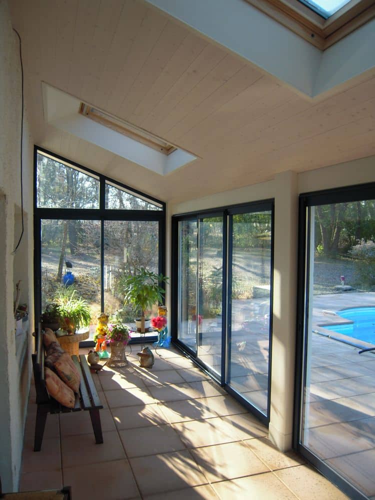 Véranda ensoleillée avec plafond lambrissé, grandes baies vitrées, sol carrelé et banquette décorée de coussins et bibelots.