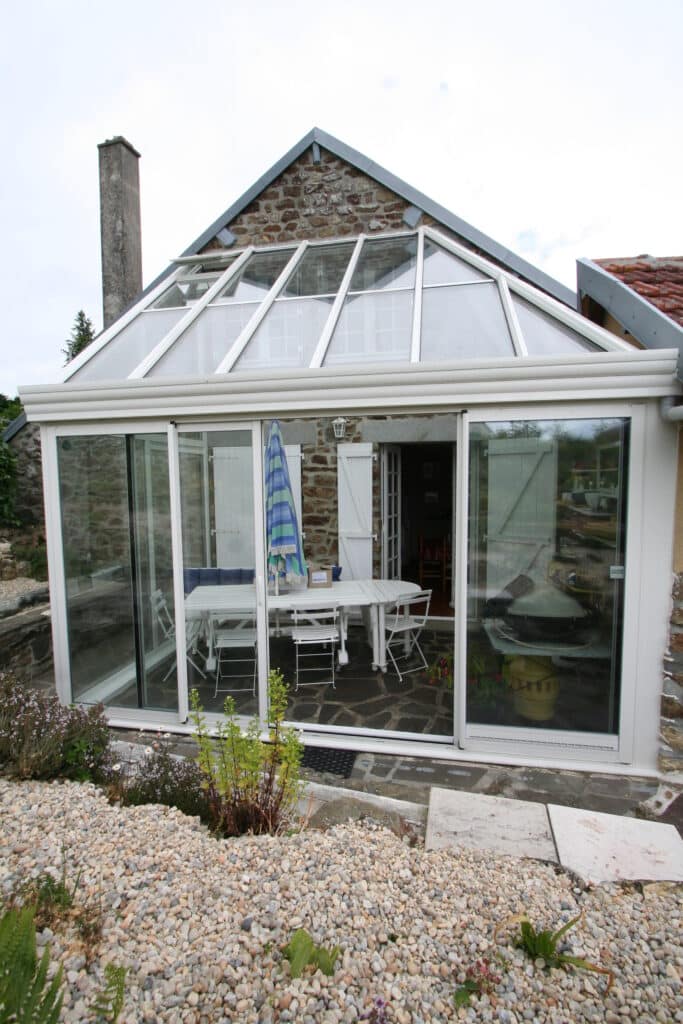 Véranda moderne vitrée à toit en verre accolée à une vieille bâtisse en pierre avec cheminée. Sol extérieur en galets et dalles.