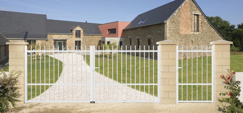 Portail et portillon blancs devant une maison en pierre avec jardin et allée de gravier.