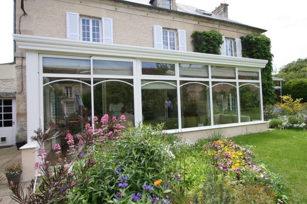Véranda blanche moderne sur une maison en pierre avec un jardin fleuri aux fleurs roses et bleues.