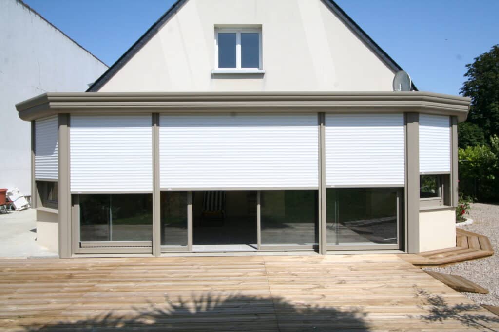 Extension de véranda beige avec volets roulants blancs et terrasse en bois devant une maison individuelle.